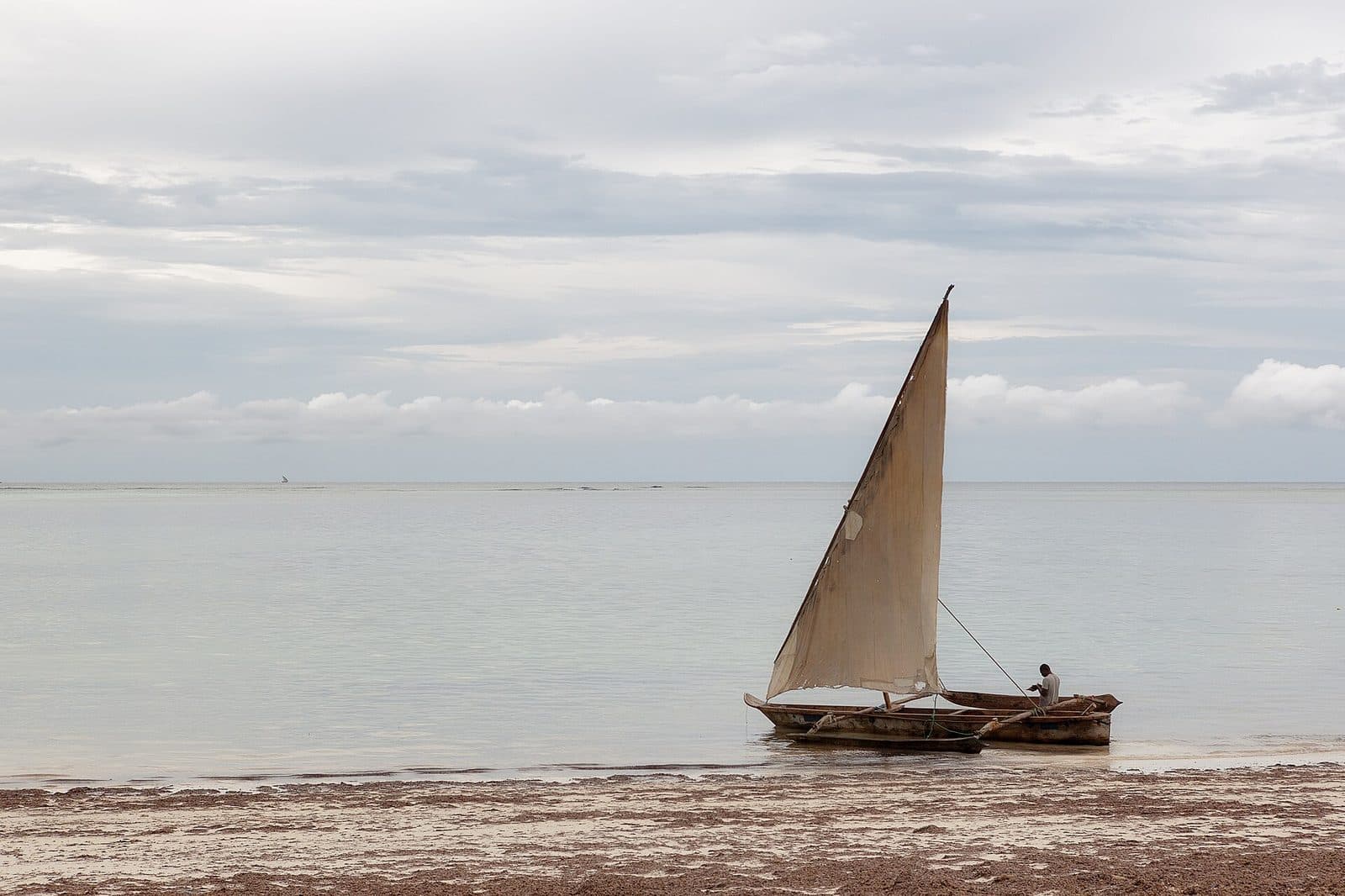 A wooden dhow with a tall sail beached on calm water under an overcast sky